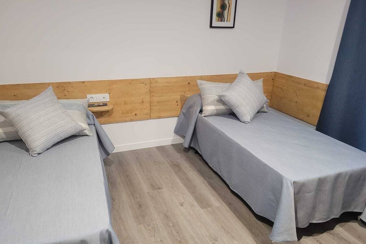 Una habitación con dos camas individuales, cada una con un almohadón a rayas y una colcha gris. La habitación tiene paneles de madera en las paredes y piso de madera.