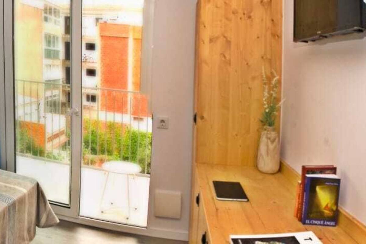 Un dormitorio con dos camas, un cabecero de madera, una mesita con un libro y vistas a un balcón con edificios.