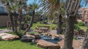 A landscaped outdoor area with palm trees, a small waterfall, and a wooden deck with seating. - Monthly Discount! Ocean View Heated Pool Hot tub! (Galveston)