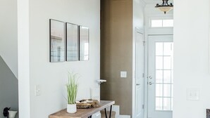 Een gang met houten vloer, een witte muur met drie ingelijste afbeeldingen, een klein houten tafeltje met een plant, en een trap die naar een andere kamer leidt.