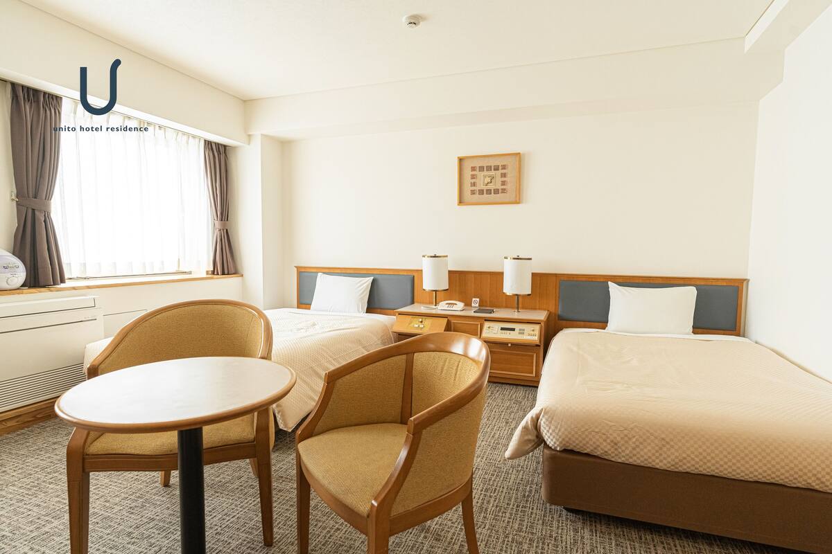 A hotel room with two beds, a small table, two chairs, a bedside table with lamps, a clock, and a framed picture on the wall.