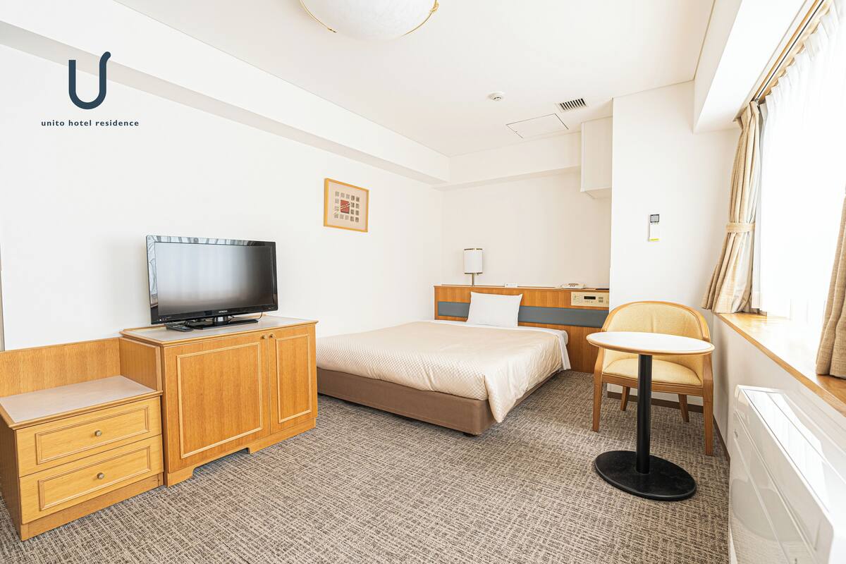 A hotel room with a bed, a television on a wooden cabinet, a small round table, a chair, and a window with curtains.