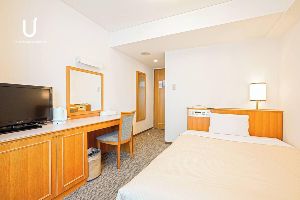A hotel room with a bed, a desk with a television, a chair, and a bedside table with a lamp.