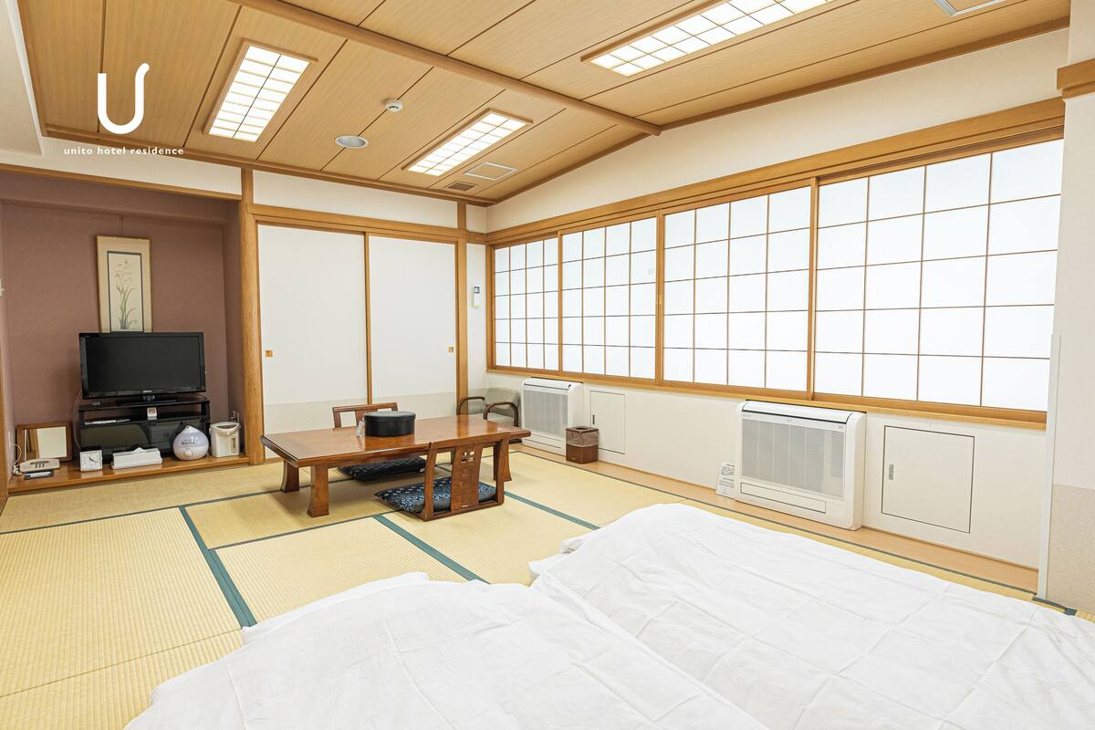 A traditional Japanese-style hotel room with tatami flooring, a futon bed, a television, a small table, and sliding doors.