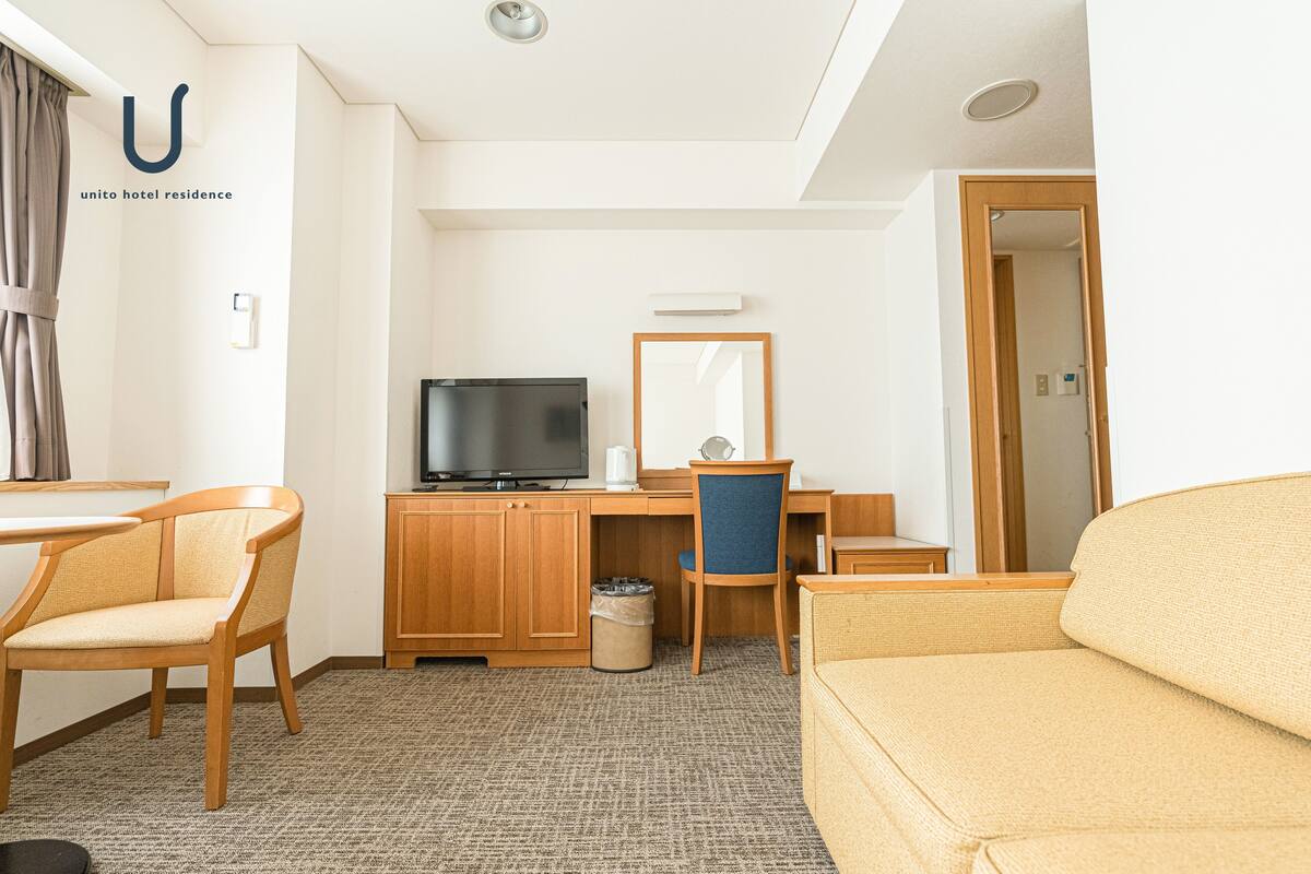 A hotel room with a wooden desk, a television, a chair, a sofa, and a mirror.