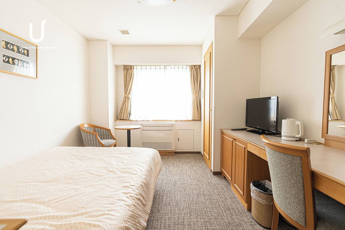 A hotel room with a bed, a desk with a television, a chair, a small table, and a window with curtains.