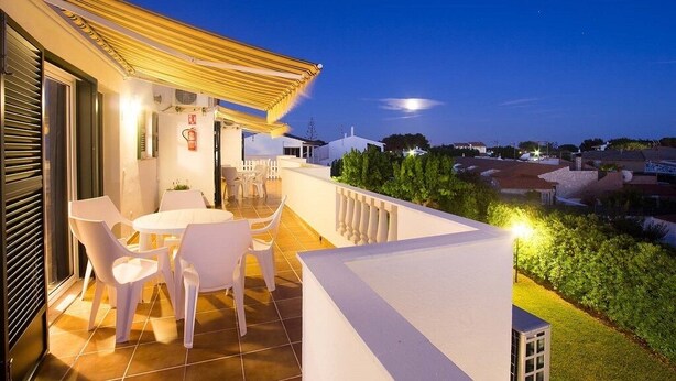 Una terraza con sillas blancas y una mesa, con vistas a un jardín y una zona residencial al atardecer.
