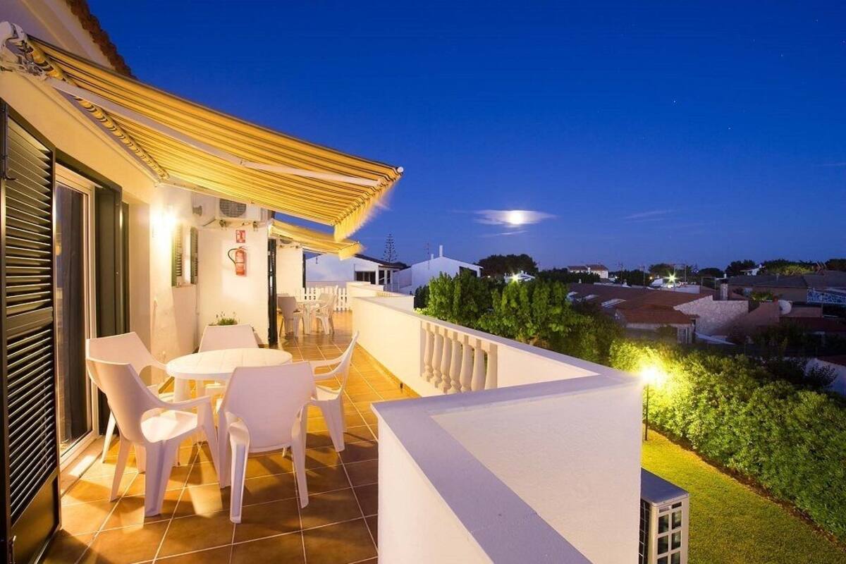 Una terraza con sillas blancas y una mesa, con vistas a un jardín y una zona residencial al atardecer.