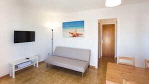 Una sala de estar minimalista con un sofá, un televisor de pantalla plana, una lámpara de pie y un cuadro de una playa.