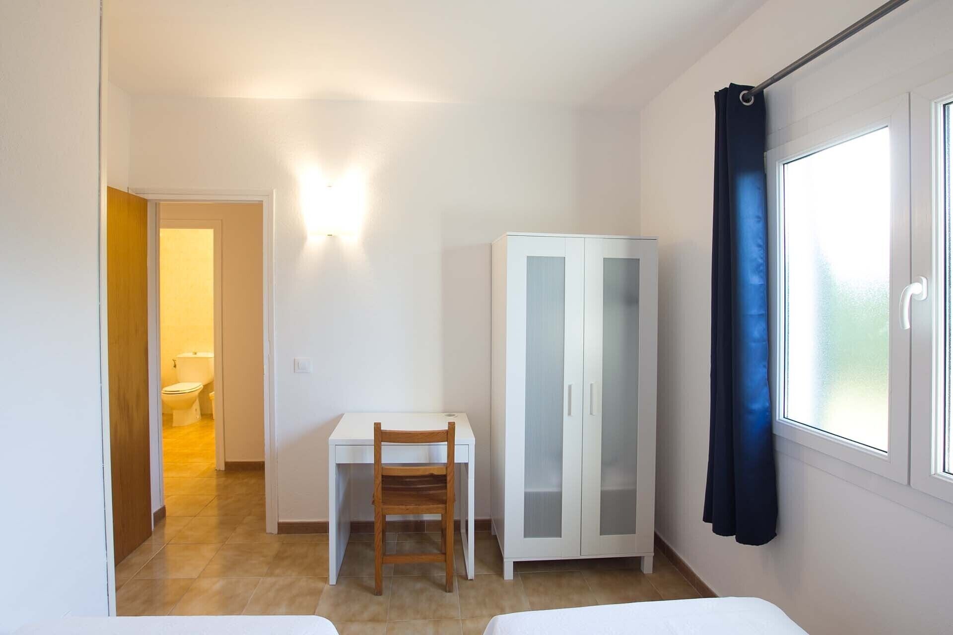 A small, well-lit room with a bed, a desk with a chair, a wardrobe, and a bathroom visible through an open door.