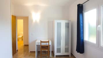 A small, well-lit room with a bed, a desk with a chair, a wardrobe, and a bathroom visible through an open door.