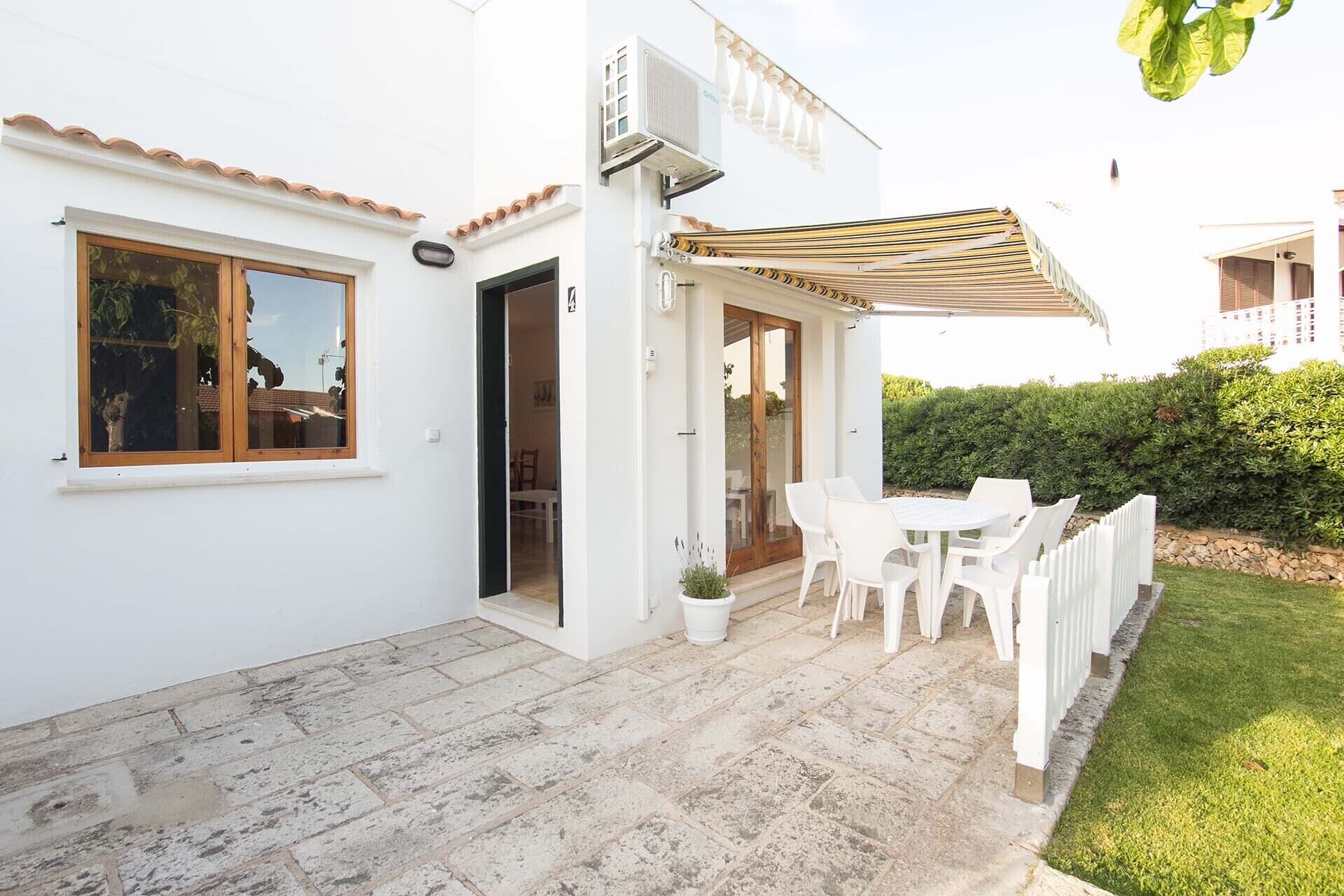 Un edificio bianco con un patio che include un tavolo, sedie, una pianta in vaso e un recinto bianco.
