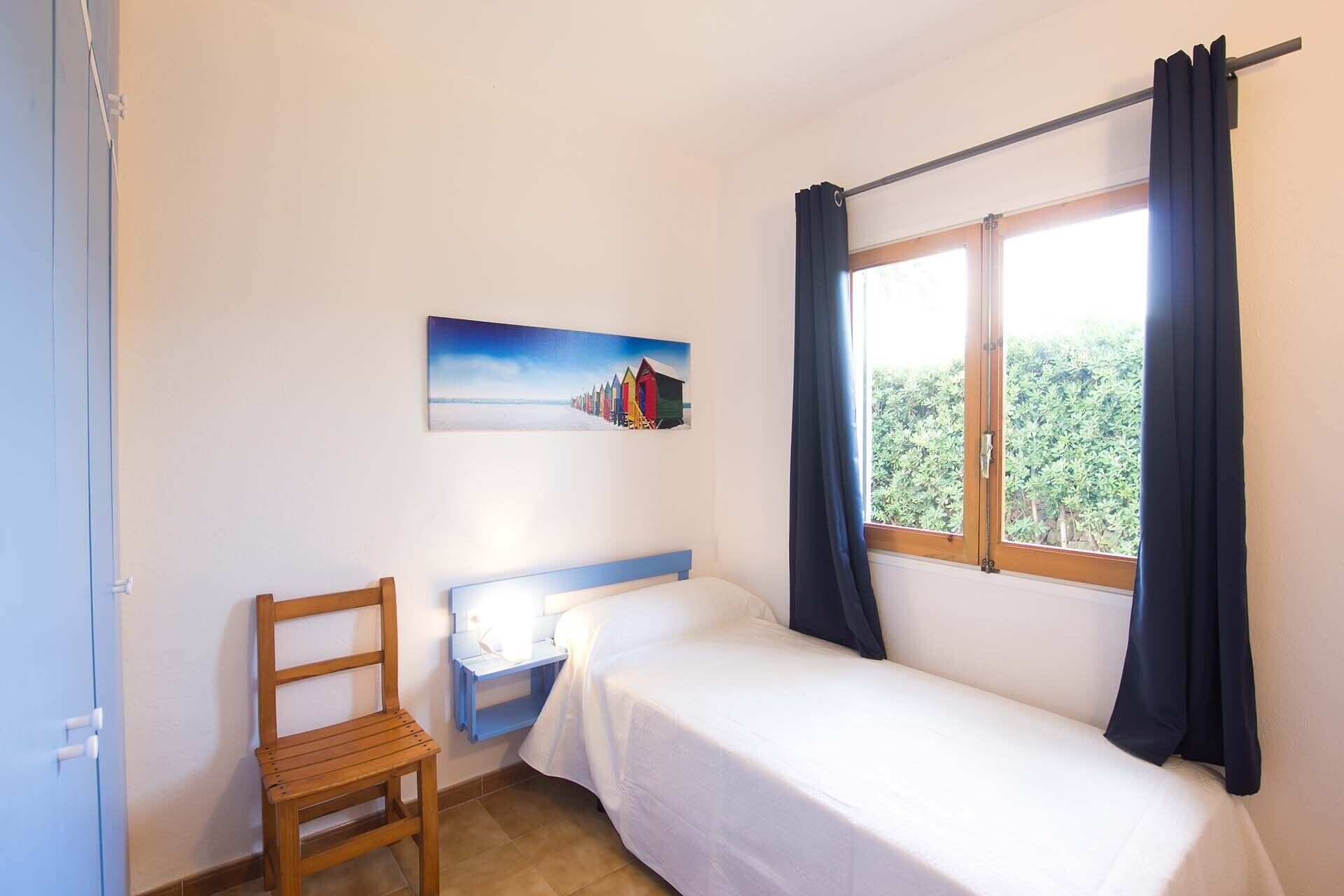 Quarto pequeno com uma cama de solteiro, uma cadeira, uma mesinha e uma janela com cortinas.