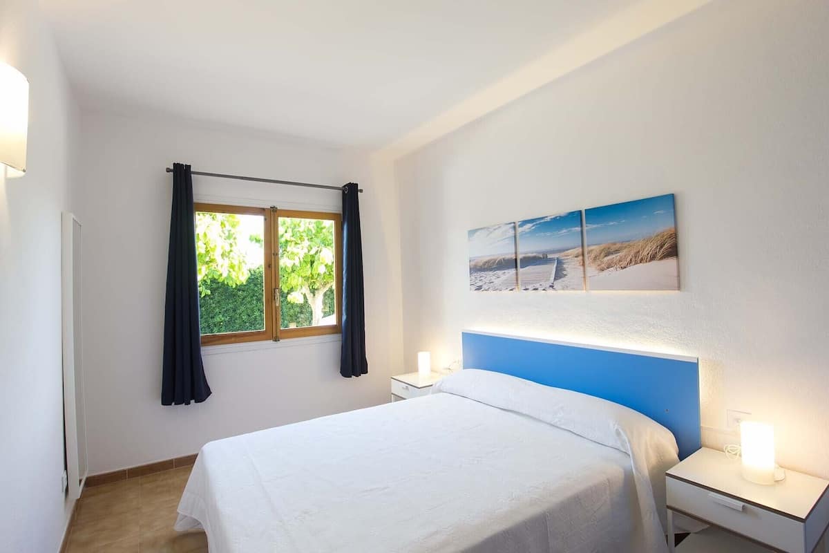 Un dormitorio con cama, mesita de noche, ventana con cortinas e imagen colgada en la pared.