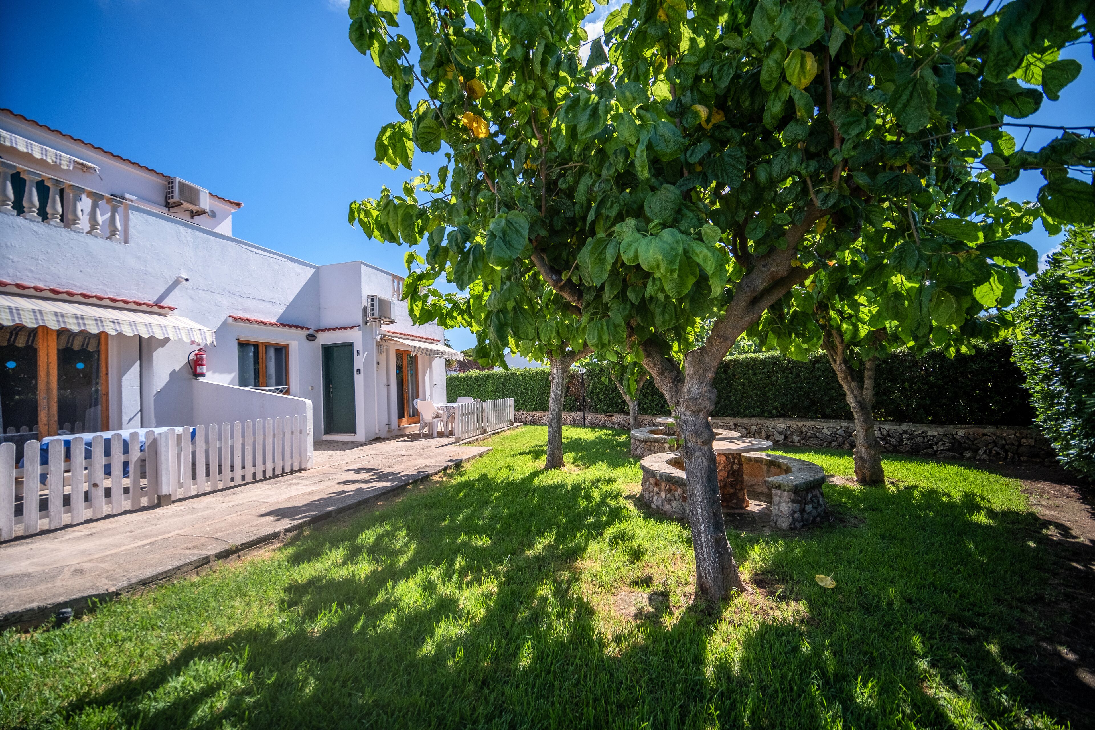 A white house with a wooden deck, a stone bench, and a tree in the yard.