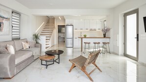 Una sala moderna con un sofá, una mesa de centro y una silla tejida original. Se ve un área de cocina con un refrigerador y taburetes de bar al fondo.