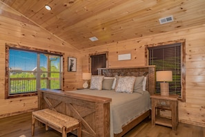 A wooden cabin bedroom with a bed, bench, bedside tables, and a view of the outdoors. - Rustic Mountain View: Mtn Views! Resort Pool, Wi-Fi, Hot Tub, Games, near Attractions (Sevierville)