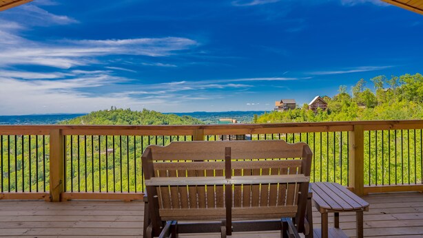 A wooden deck with a bench and table, overlooking a lush green landscape and distant buildings. - 🏔️Rustic Mountain View🌲Resort Pool, Hot Tub & Game Room (Sevierville)