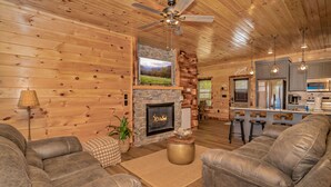 A cozy log cabin interior with a stone fireplace, leather sofas, a kitchen area, and a ceiling fan. - 🏔️Rustic Mountain View🌲Resort Pool, Hot Tub & Game Room (Sevierville)