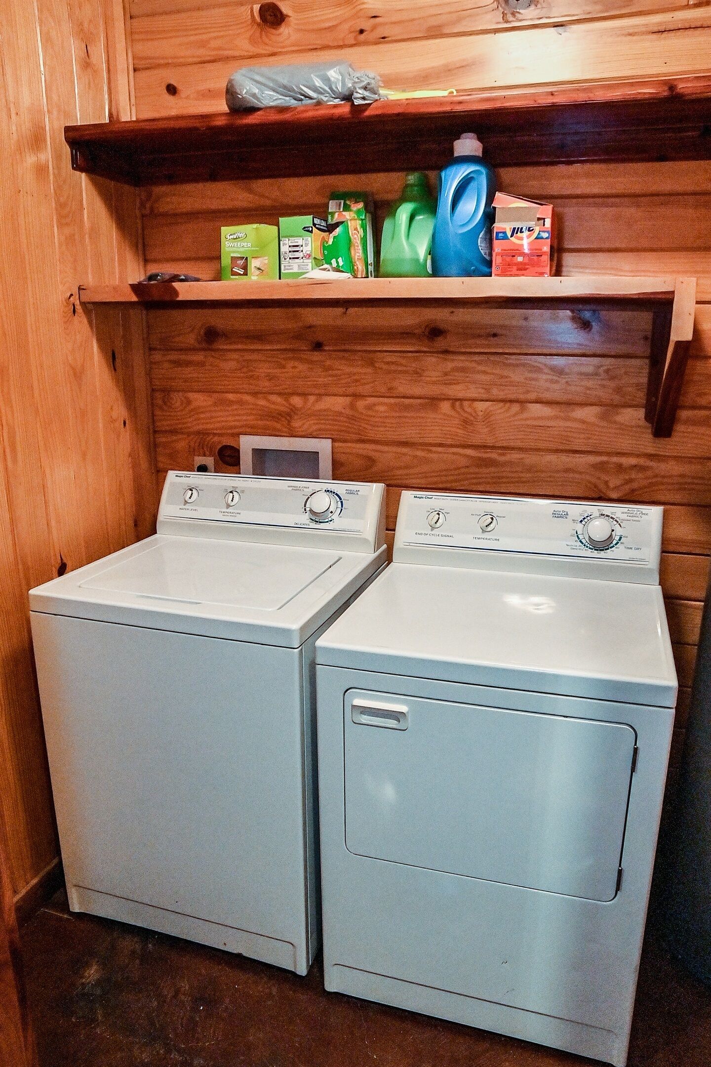 A laundry area with two white front-loading washers and dryers, wooden shelves above holding cleaning supplies, and a wooden wall.