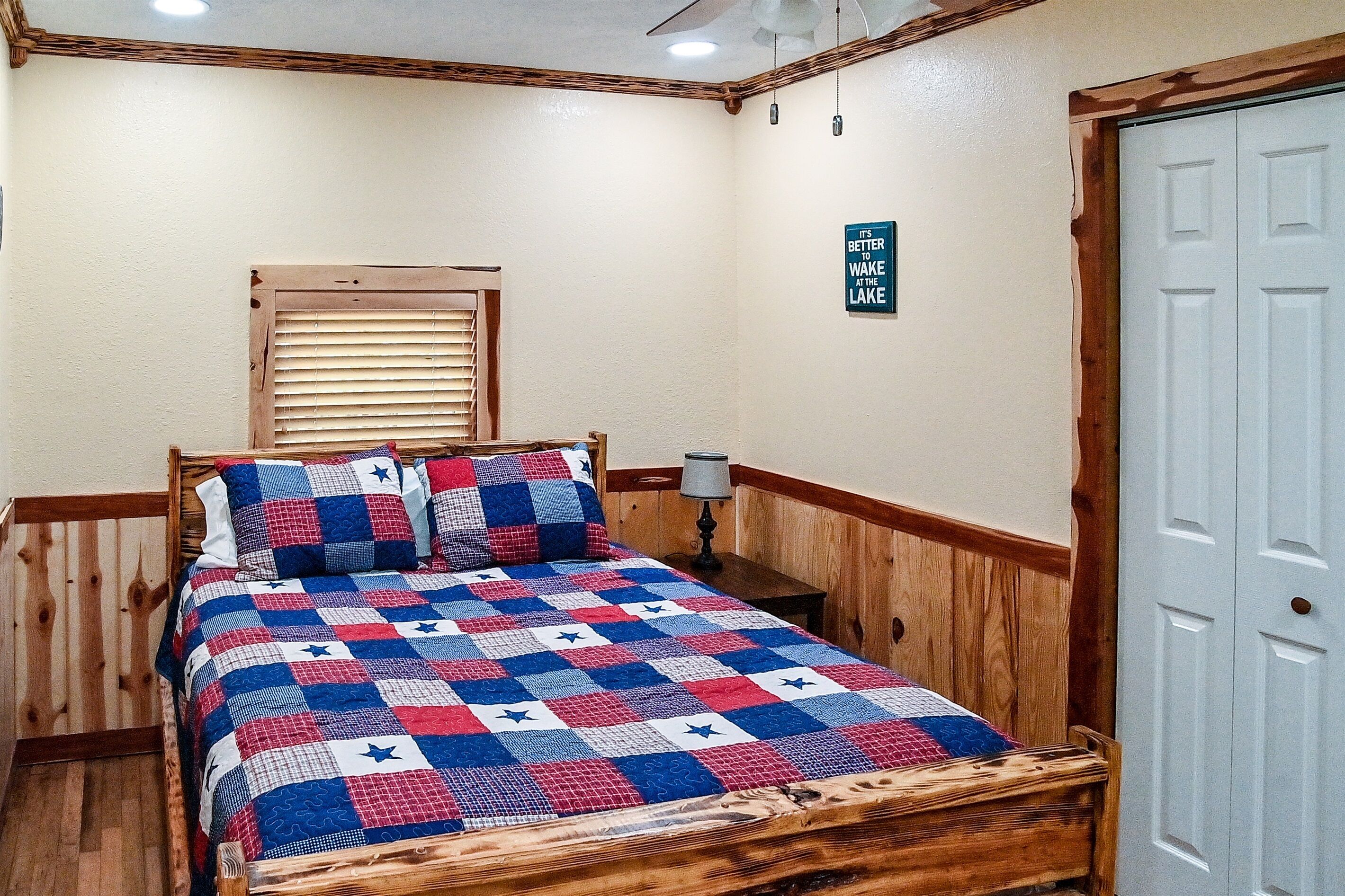 A bedroom with a wooden bed, a window with blinds, a nightstand with a lamp, and a door with a sign.