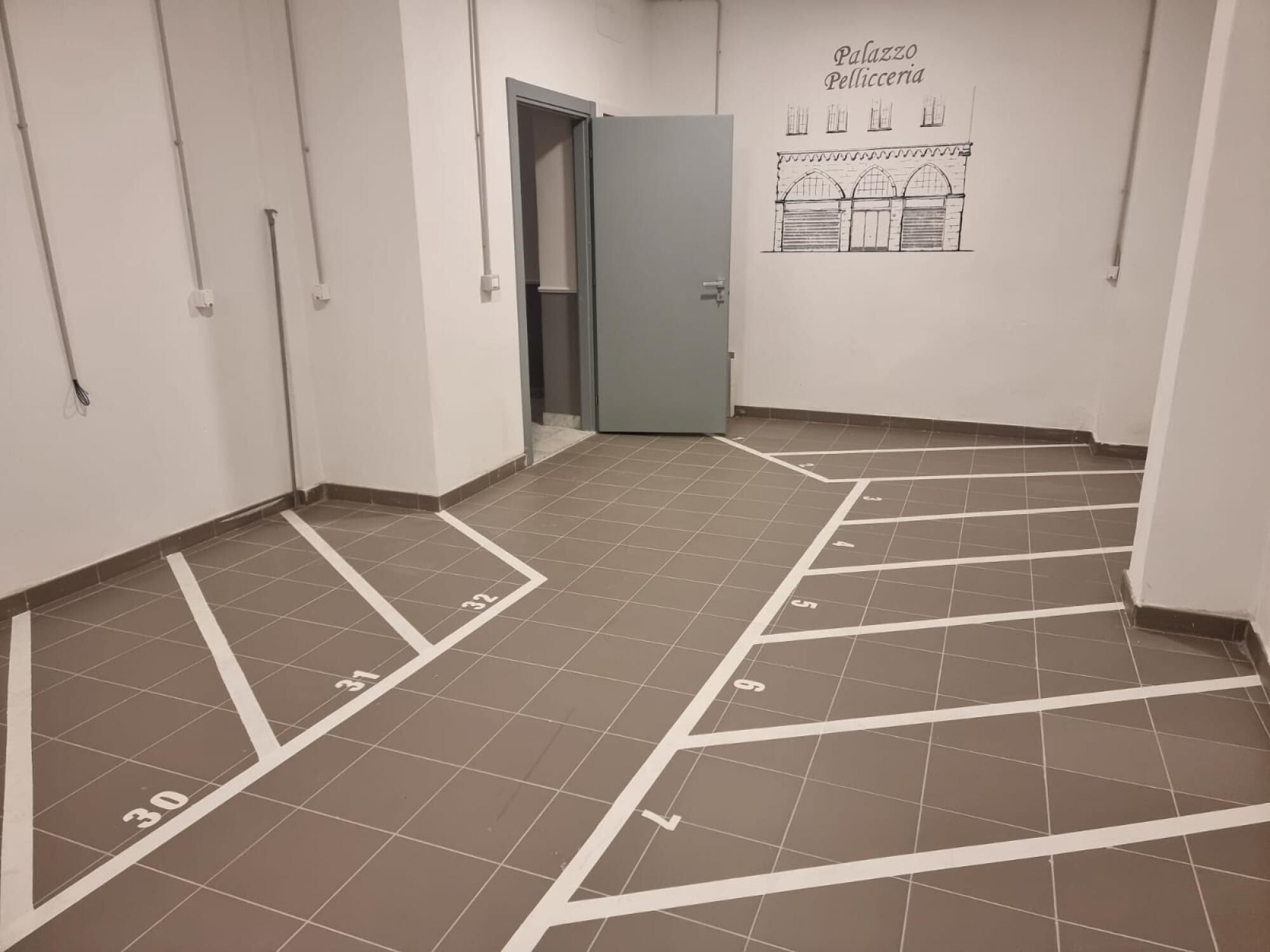 Käytävällä on numeroituja parkkipaikkoja, ovi ja seinä, jossa on piirros ja teksti: ”Palazzo Pelliceria”.