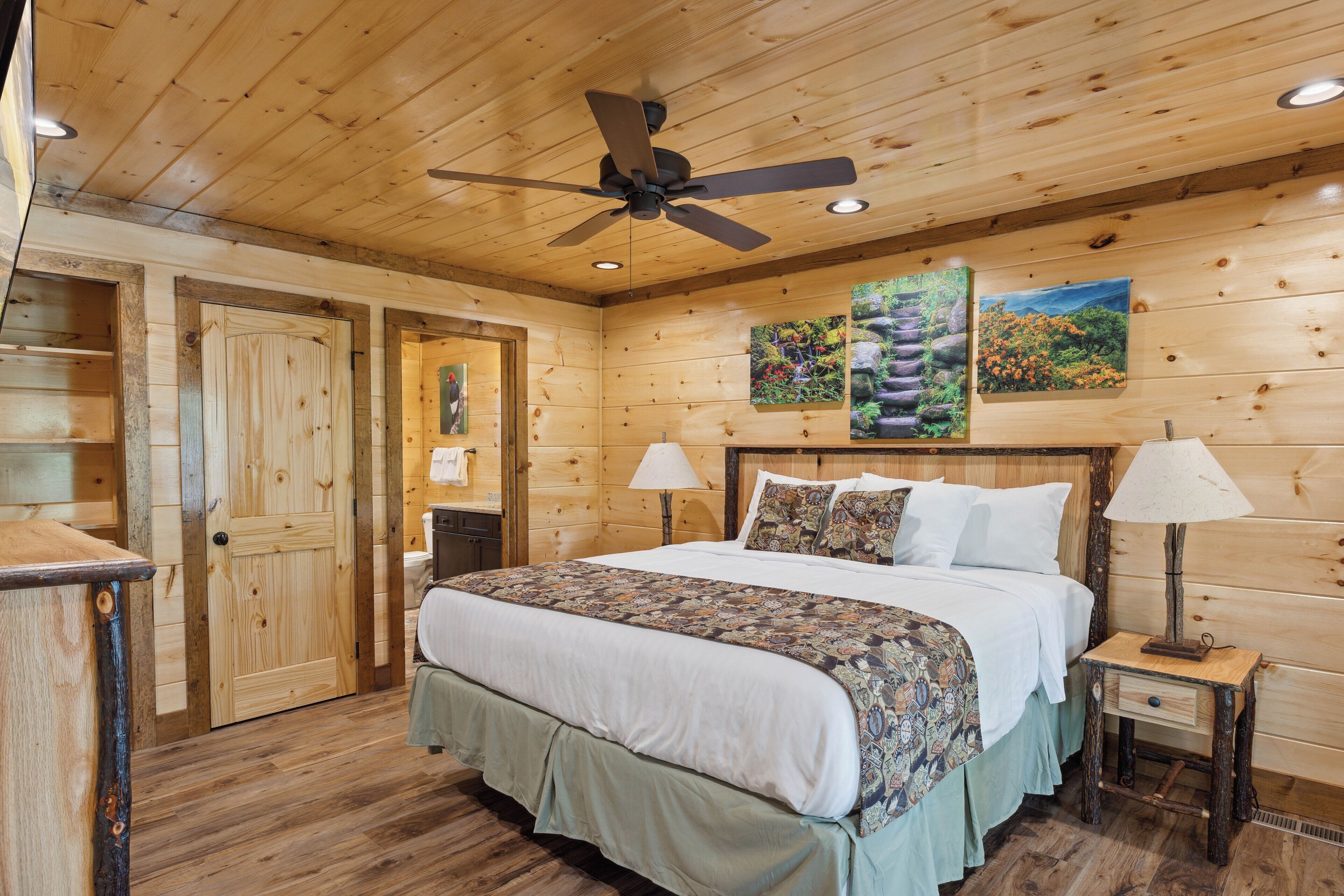 A wooden cabin bedroom with a bed, two bedside tables, a ceiling fan, and two paintings on the wall.