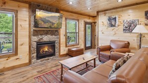 L’intérieur chaleureux d’un chalet avec une cheminée en pierre, des meubles en cuir et de grandes fenêtres offrant une vue sur le paysage extérieur.