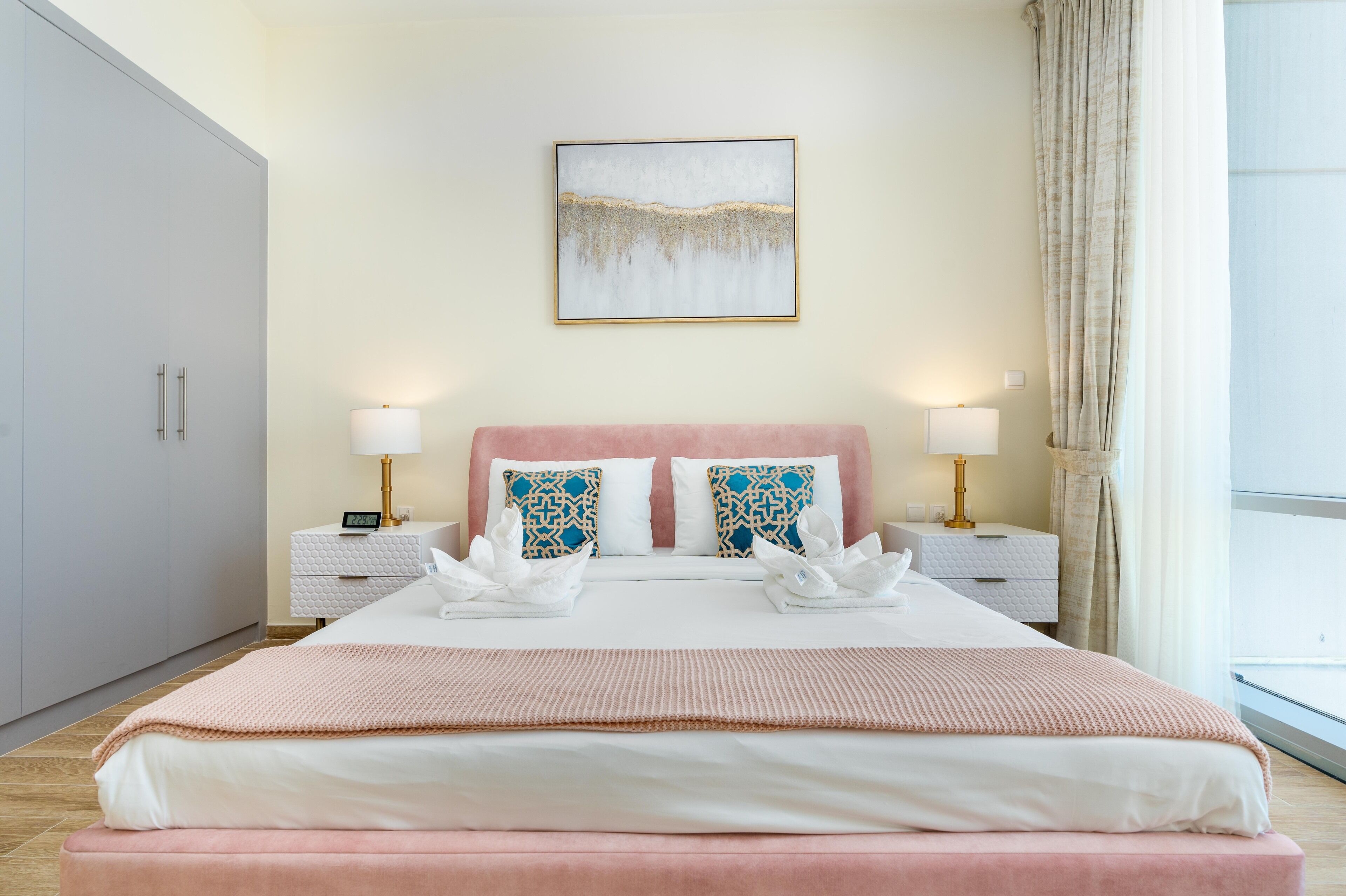 A neatly made bed with a pink headboard, two bedside tables with lamps, a wall-mounted artwork, and a large window with curtains.