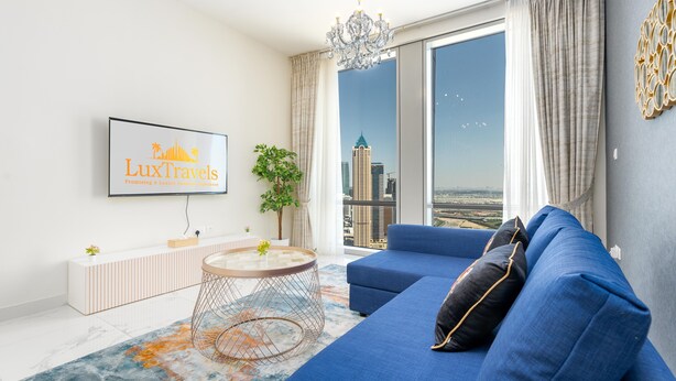 A modern living room with a large sofa, a TV mounted on the wall, and a view of a cityscape through a large window. - Lux BnB I Amna Tower I Panaromic Views (Dubai)