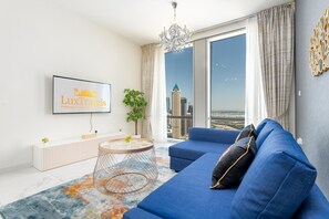 A modern living room with a large sofa, a TV mounted on the wall, and a view of a cityscape through a large window. - Lux BnB I Amna Tower I Panaromic Views (Dubai)