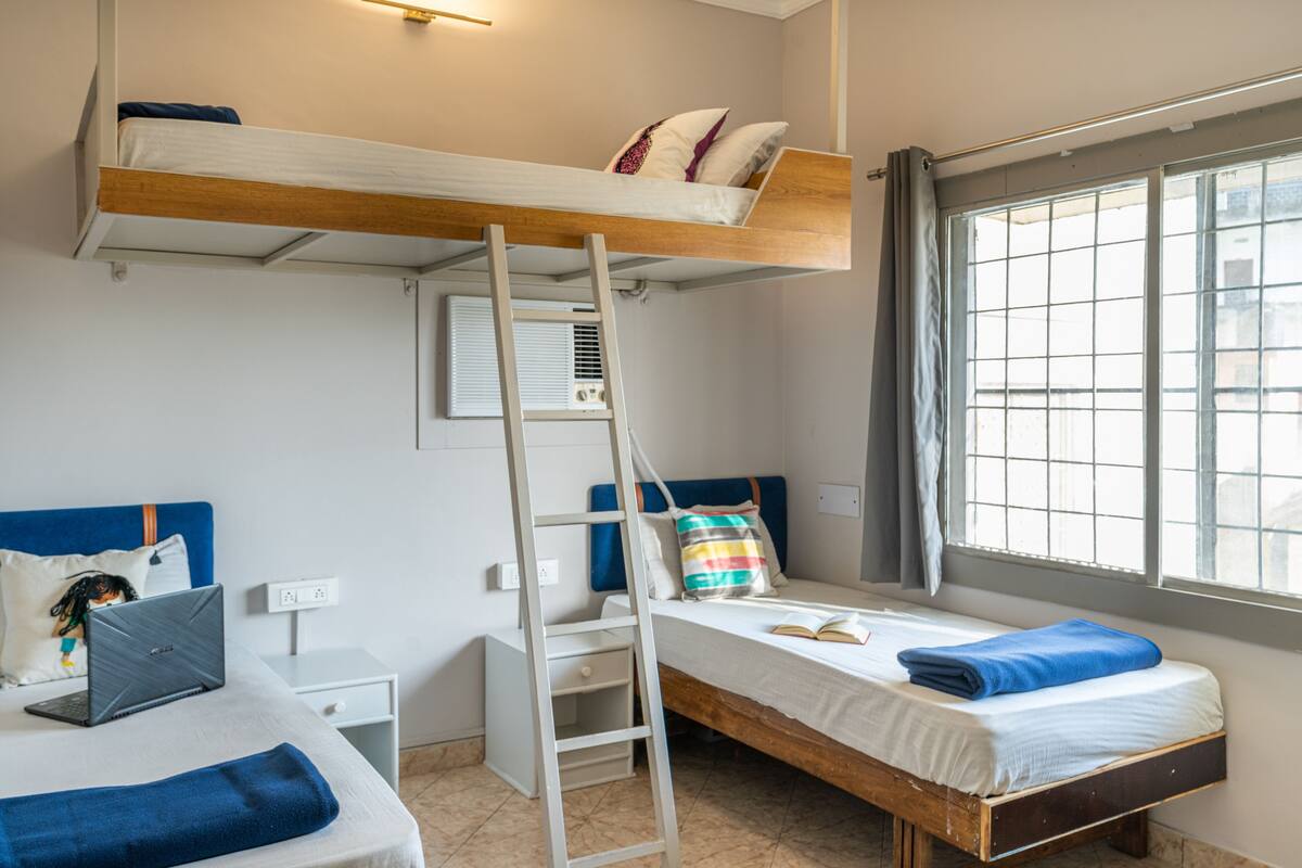 A dorm-style room with bunk beds, a ladder, a desk with a laptop, and a window with blinds.