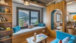 A cozy living room with a large window, wooden shelves, a blue sofa, and a small table with a jigsaw puzzle. - Madpackers Rishikesh (Yamkeshwar)