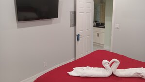 Een slaapkamer met een rode sprei, een flatscreen televisie aan de muur en een witte deur naar een badkamer.