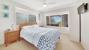 Une chambre à coucher comprenant un lit, une table de chevet, une lampe et un ventilateur de plafond. Il y a des fenêtres avec des stores et un téléviseur fixé au mur.