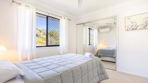 Un dormitorio con cama, ventana con cortinas, espejo y un cuadro en la pared.