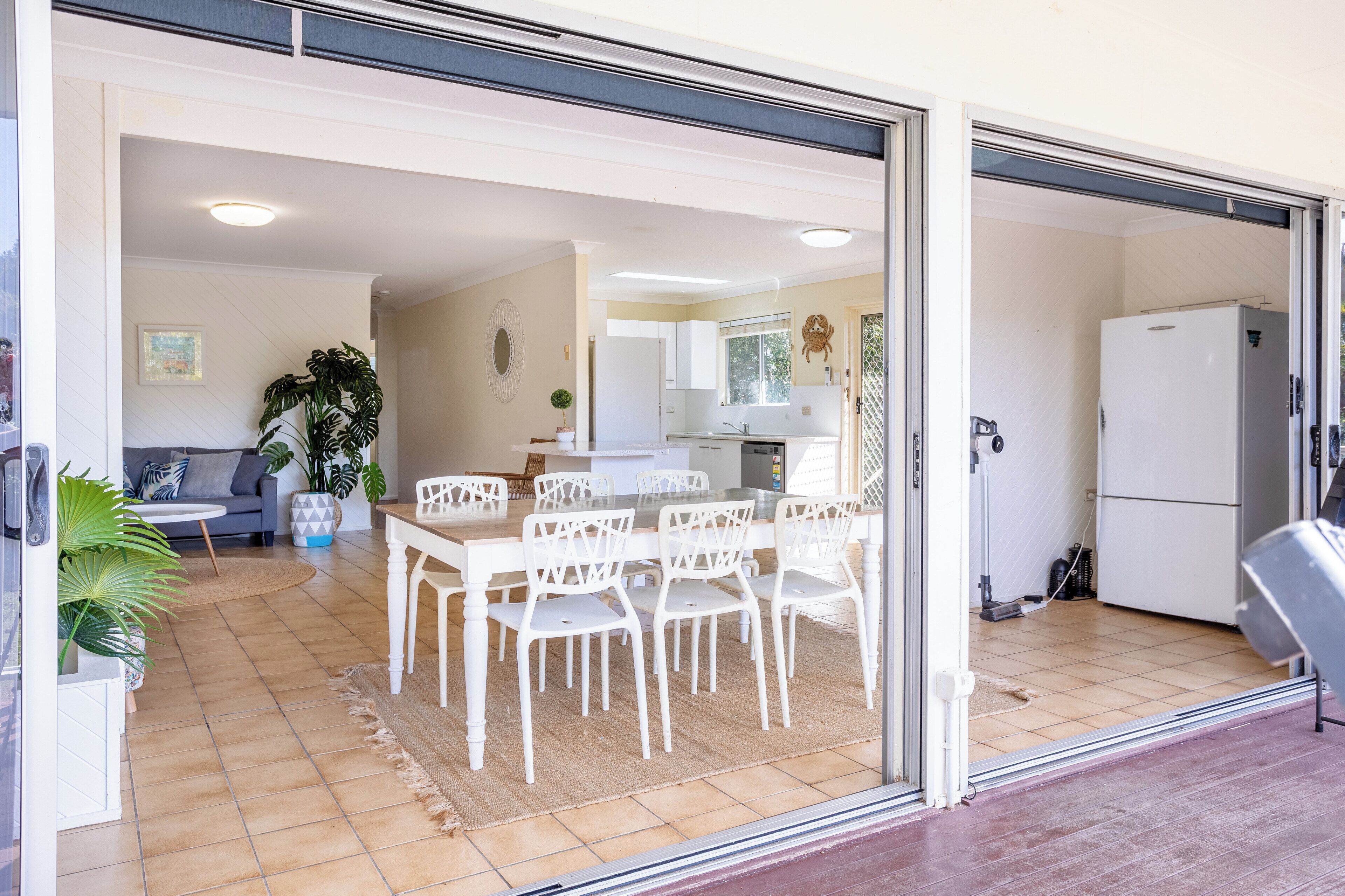 A dining area with a white table and chairs, a kitchen with a refrigerator, and a living room with a sofa and plants.