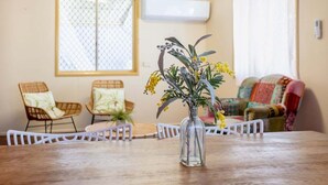 A dining area with a wooden table, white chairs, a glass vase with flowers, wicker chairs, a colorful sofa, and a window with a patterned blind. - The Normanby - For the traveller on a budget (Yeppoon)
