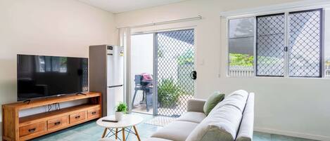A living room with a flat-screen TV, wooden entertainment unit, sofa, and a sliding glass door leading to an outdoor grill area.