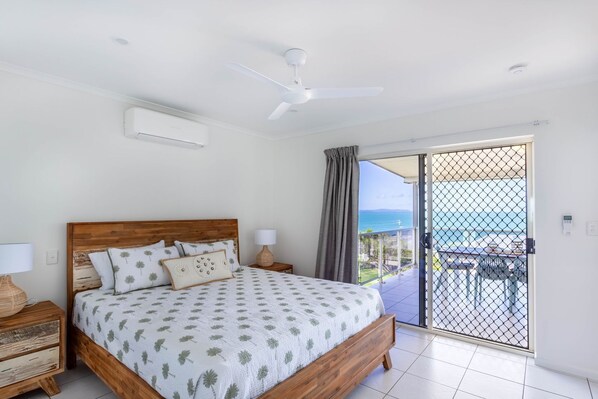 A bedroom with a wooden bed, two bedside tables with lamps, a ceiling fan, and a view of the sea through a balcony door. - Catalina Island Views - What an understatement! (Yeppoon)