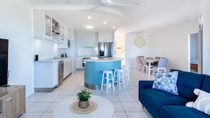 A modern kitchen with white cabinets, a central island with stools, a dining area with a table and chairs, and a living room with a blue sofa and a TV. - Catalina Island Views - What an understatement! (Yeppoon)