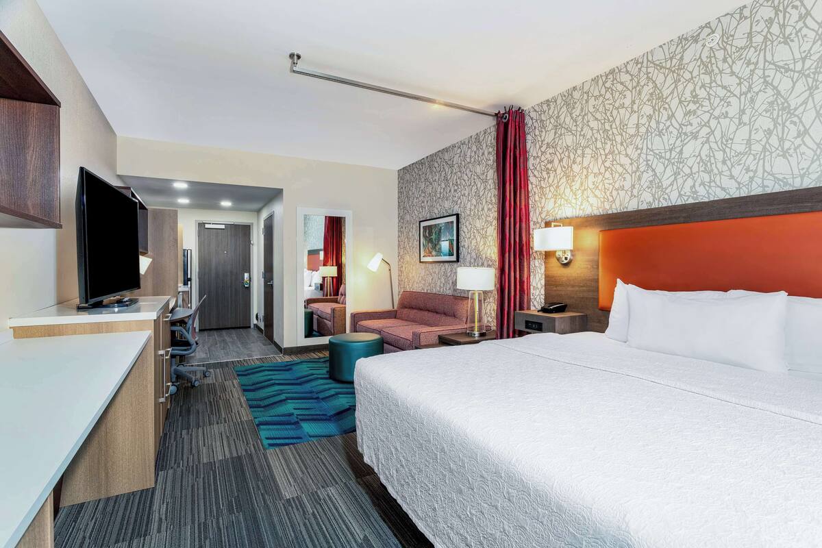 A hotel room with a bed, a desk with a TV, a chair, a sofa, and a bathroom visible through an open door.