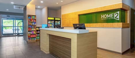 A hotel reception desk with a sign reading "HOME 2 SUITES BY HILTON," a computer, and a shelf with snacks.