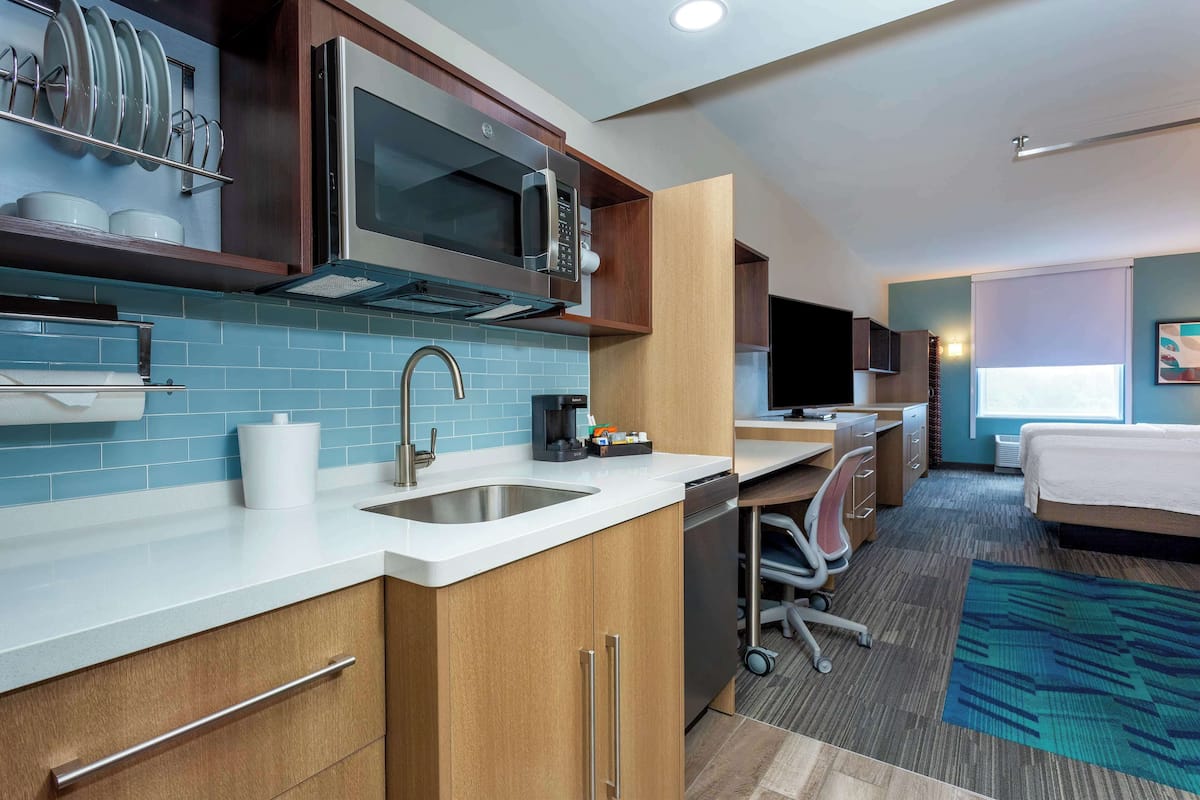 A compact hotel room with a kitchenette, a bed, a desk, and a large window.