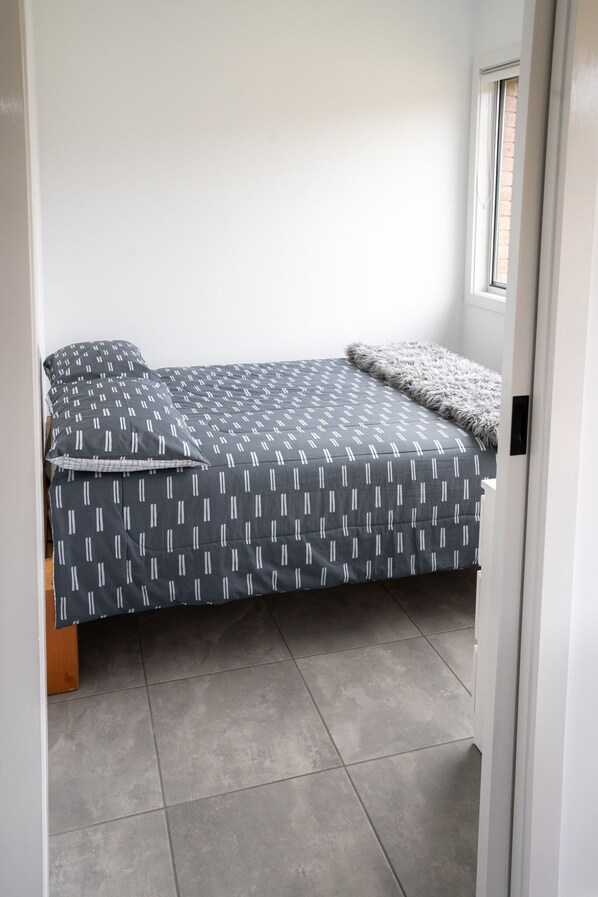 Una habitación pequeña con cama individual, una colcha gris estampada, un plaid gris mullido y una ventana con vista a un muro de ladrillo.