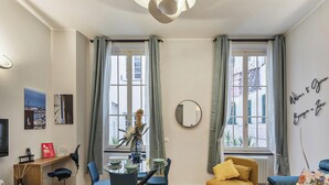 Ein modernes Wohnzimmer mit Essbereich, großen Fenstern mit Vorhängen, einem runden Spiegel und einer Couch.