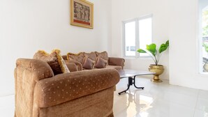 Un salón con un sofá marrón, una mesa de centro de vidrio, un cuadro enmarcado en la pared y una planta en maceta cerca de la ventana.