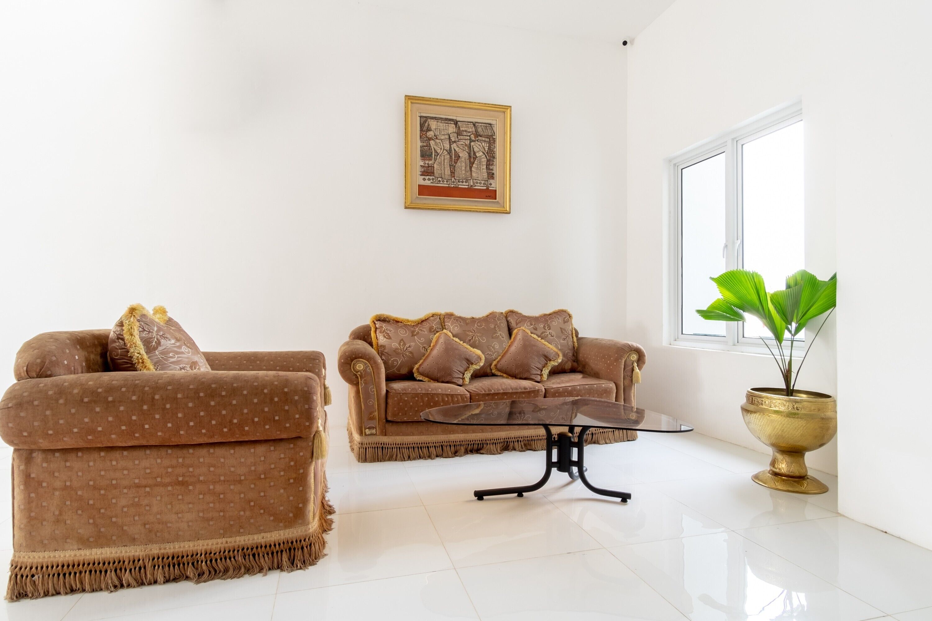 A living room with two brown sofas, a glass coffee table, a framed artwork on the wall, and a potted plant by the window.