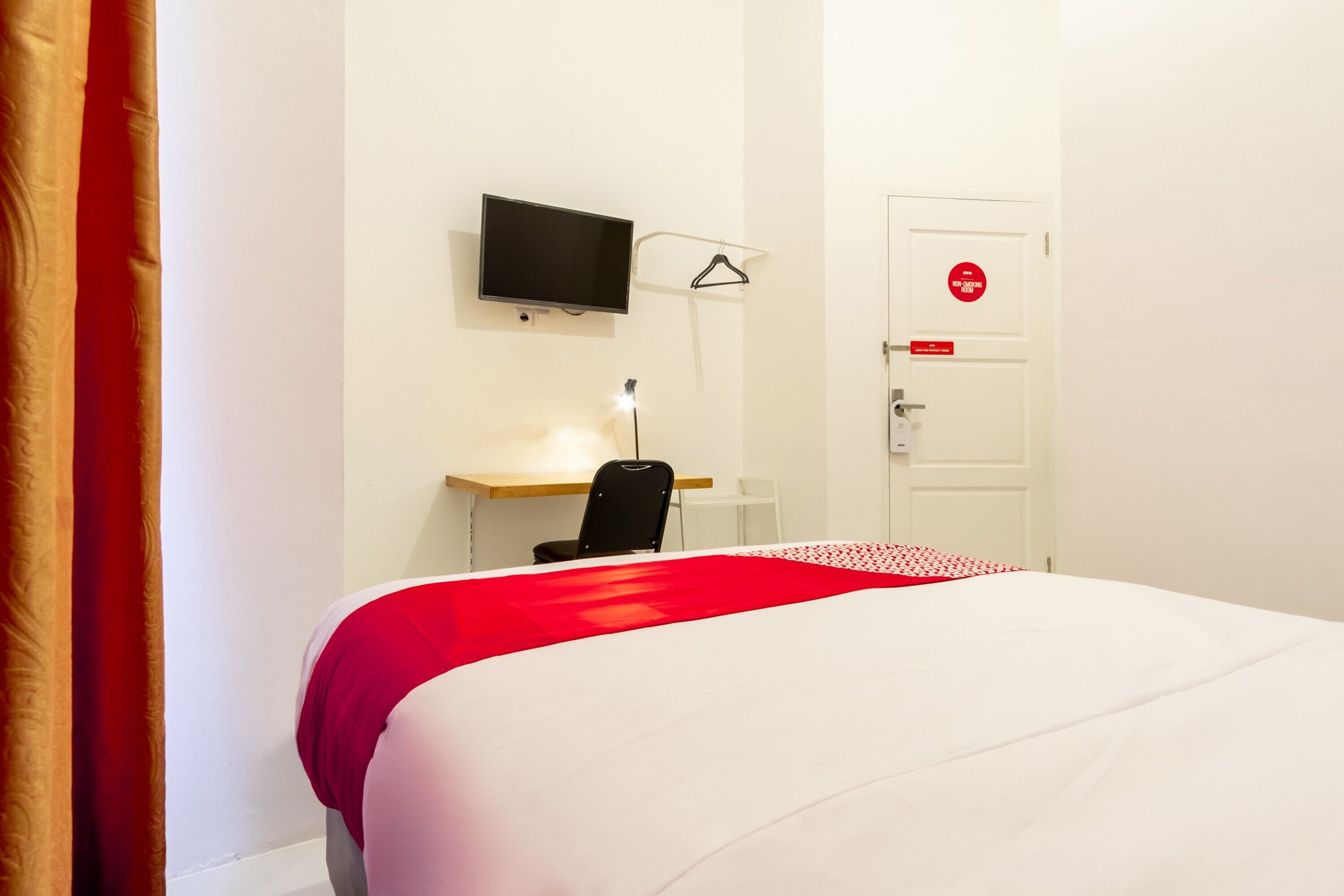Una habitación de hotel con cama, escritorio con silla, televisor montado en la pared y una puerta con letrero rojo.