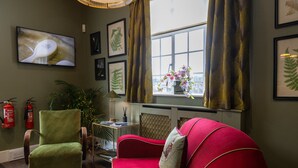A room with a green armchair, a red sofa, a television, framed botanical prints, a window with curtains, and a fire extinguisher. - Maidens Tower B&B (Maidstone)